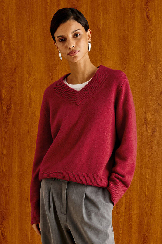 A woman wearing a burgundy V-neck knit sweater over a white undershirt and gray trousers, standing against a warm wood-paneled background.