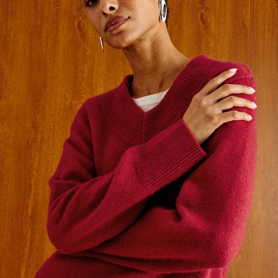 A woman wearing a deep burgundy knit V-neck sweater over a white undershirt and gray trousers, posed against a warm wood-paneled background.