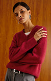 A woman wearing a deep burgundy knit V-neck sweater over a white undershirt and gray trousers, posed against a warm wood-paneled background.