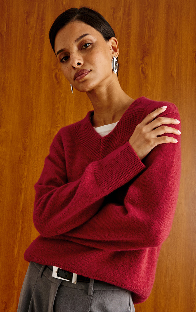 A woman wearing a deep burgundy knit V-neck sweater over a white undershirt and gray trousers, posed against a warm wood-paneled background.