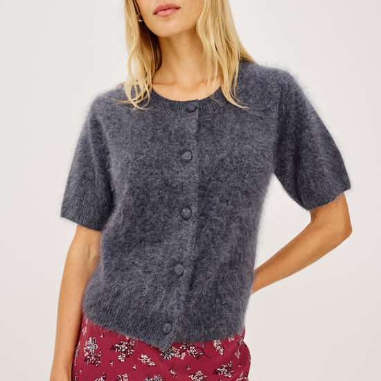 A woman modeling a short-sleeve fuzzy knit cardigan in a heathered slate blue-gray color, paired with a red floral skirt, against a plain light gray studio background.