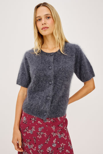 A woman modeling a short-sleeve fuzzy knit cardigan in a heathered slate blue-gray color, paired with a red floral skirt, against a plain light gray studio background.