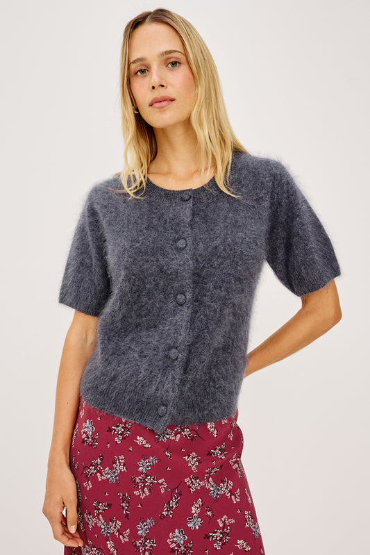 A woman modeling a short-sleeve fuzzy knit cardigan in a heathered slate blue-gray color, paired with a red floral skirt, against a plain light gray studio background.