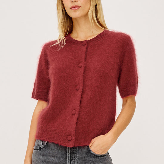 A woman modeling a short-sleeved, fuzzy knit cardigan in a deep burgundy red, paired with gray jeans, against a plain light gray studio background.