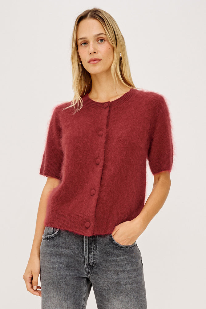 A woman modeling a short-sleeved, fuzzy knit cardigan in a deep burgundy red, paired with gray jeans, against a plain light gray studio background.