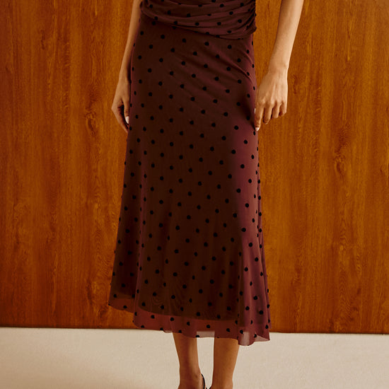 A woman models a sleeveless, ruched midi dress in deep burgundy with black polka dots, paired with black strappy sandals and a small black handbag, set against a warm wood-paneled wall.