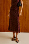 A woman models a sleeveless, ruched midi dress in deep burgundy with black polka dots, paired with black strappy sandals and a small black handbag, set against a warm wood-paneled wall.