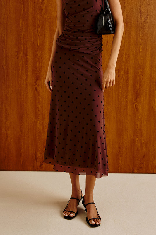 A woman models a sleeveless, ruched midi dress in deep burgundy with black polka dots, paired with black strappy sandals and a small black handbag, set against a warm wood-paneled wall.
