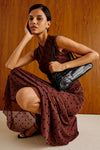 A woman crouches in a studio setting against a warm wood-paneled wall, wearing a sleeveless rust-brown dress with black polka dots and carrying a black crocodile-embossed shoulder bag.