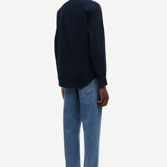 A man shown from the back wearing a dark navy long-sleeve button-up shirt, light blue jeans, and black leather shoes against a plain light gray background.