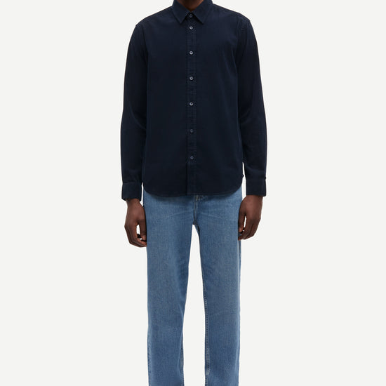 A man modeling a dark navy blue long-sleeve button-up shirt with light blue jeans and black shoes, standing against a plain light gray background.