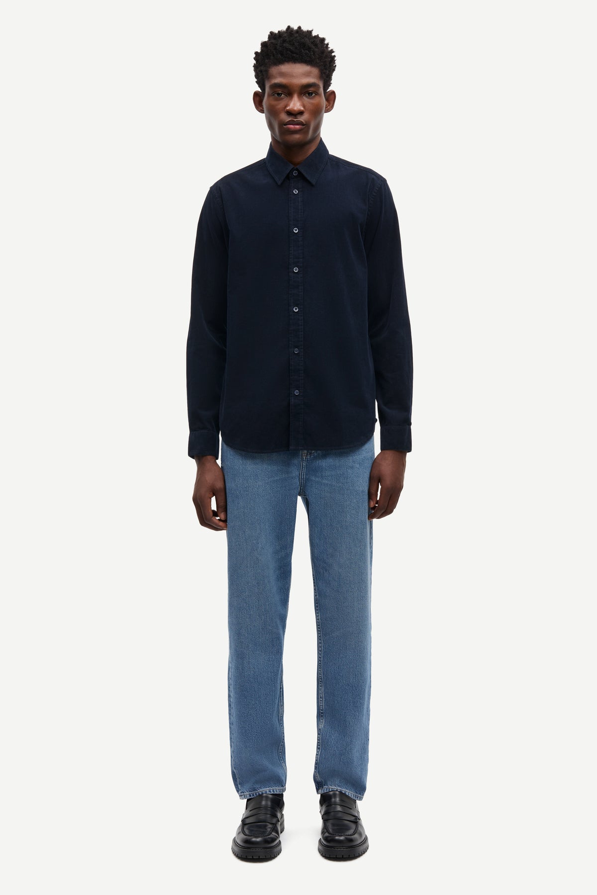 A man modeling a dark navy blue long-sleeve button-up shirt with light blue jeans and black shoes, standing against a plain light gray background.