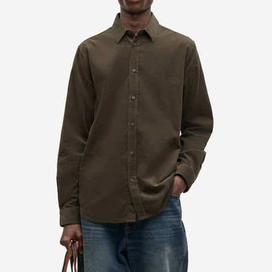 A man wearing a dark olive brown long-sleeve button-down shirt and blue jeans, holding a brown leather tote bag against a light gray background.