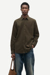 A man wearing a dark olive brown long-sleeve button-down shirt and blue jeans, holding a brown leather tote bag against a light gray background.