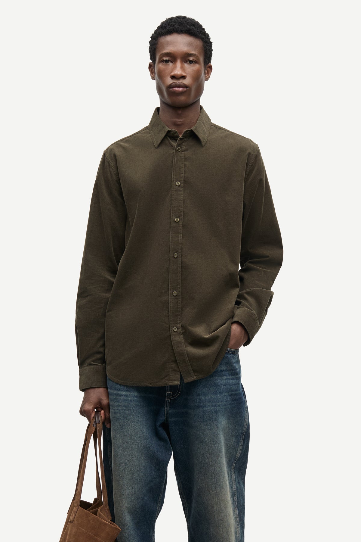 A man wearing a dark olive brown long-sleeve button-down shirt and blue jeans, holding a brown leather tote bag against a light gray background.