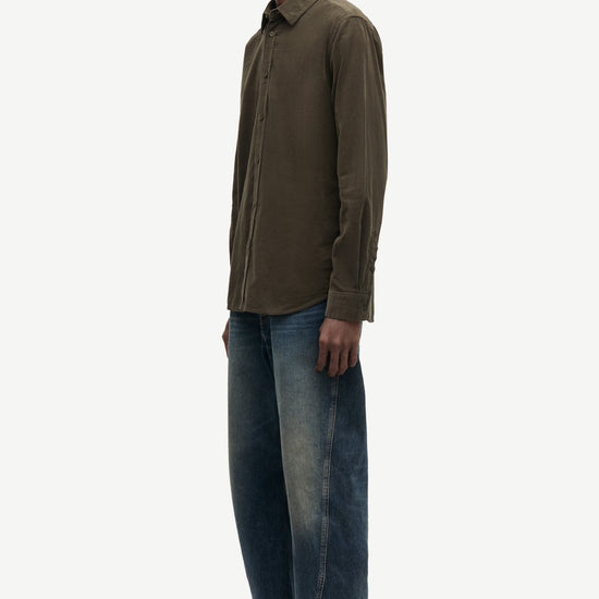 A man shown in profile wearing a dark olive brown long-sleeve button-up shirt, blue jeans, and brown shoes against a plain light gray studio background.
