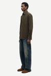 A man shown in profile wearing a dark olive brown long-sleeve button-up shirt, blue jeans, and brown shoes against a plain light gray studio background.