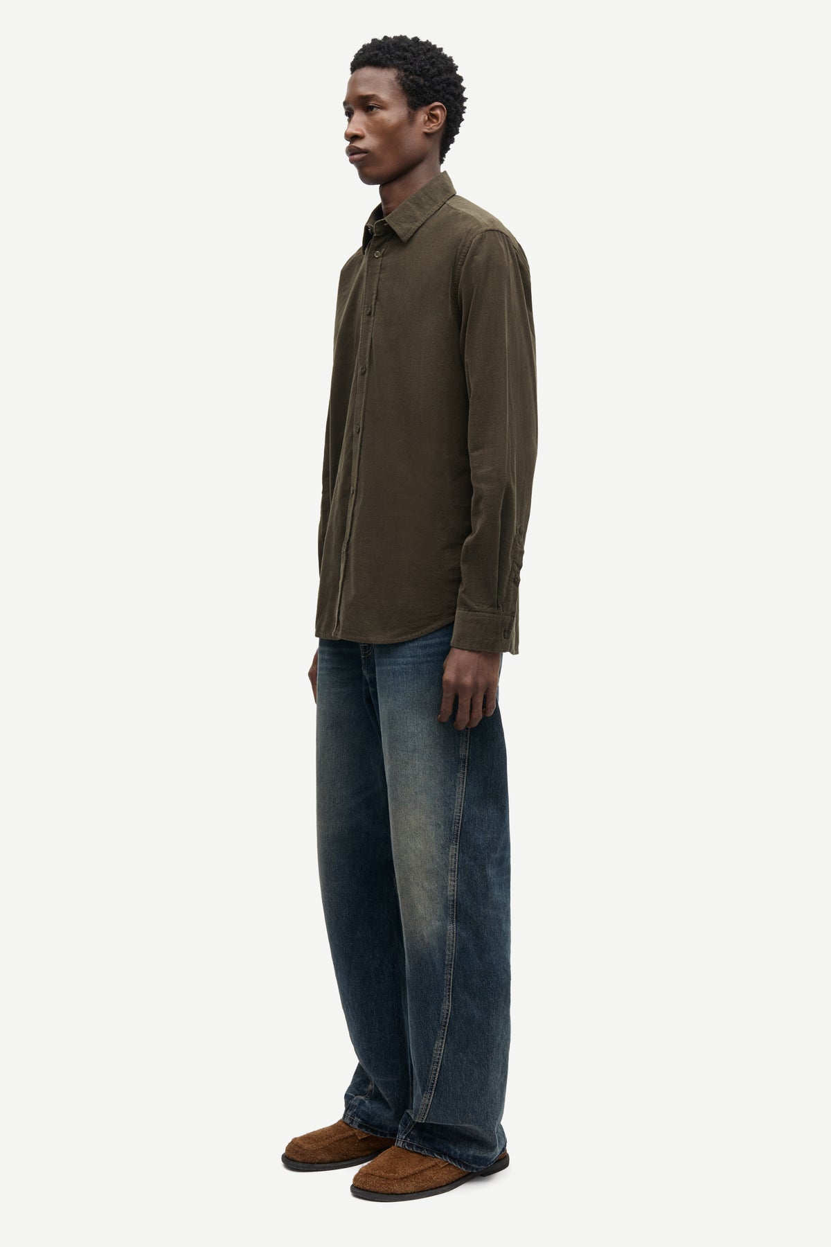 A man shown in profile wearing a dark olive brown long-sleeve button-up shirt, blue jeans, and brown shoes against a plain light gray studio background.