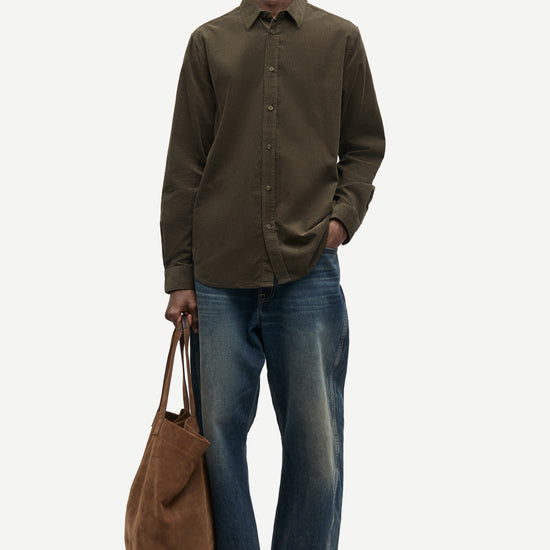A man modeling a dark olive green long-sleeve button-up shirt, blue jeans, and brown suede shoes, holding a brown canvas tote bag against a light gray studio background.