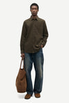 A man modeling a dark olive green long-sleeve button-up shirt, blue jeans, and brown suede shoes, holding a brown canvas tote bag against a light gray studio background.
