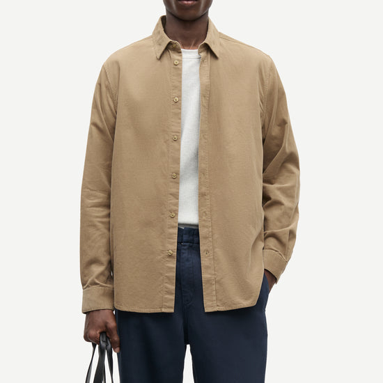 A man modeling a tan long-sleeve button-up shirt over a white t-shirt and navy blue trousers, holding a black tote bag against a light gray background.