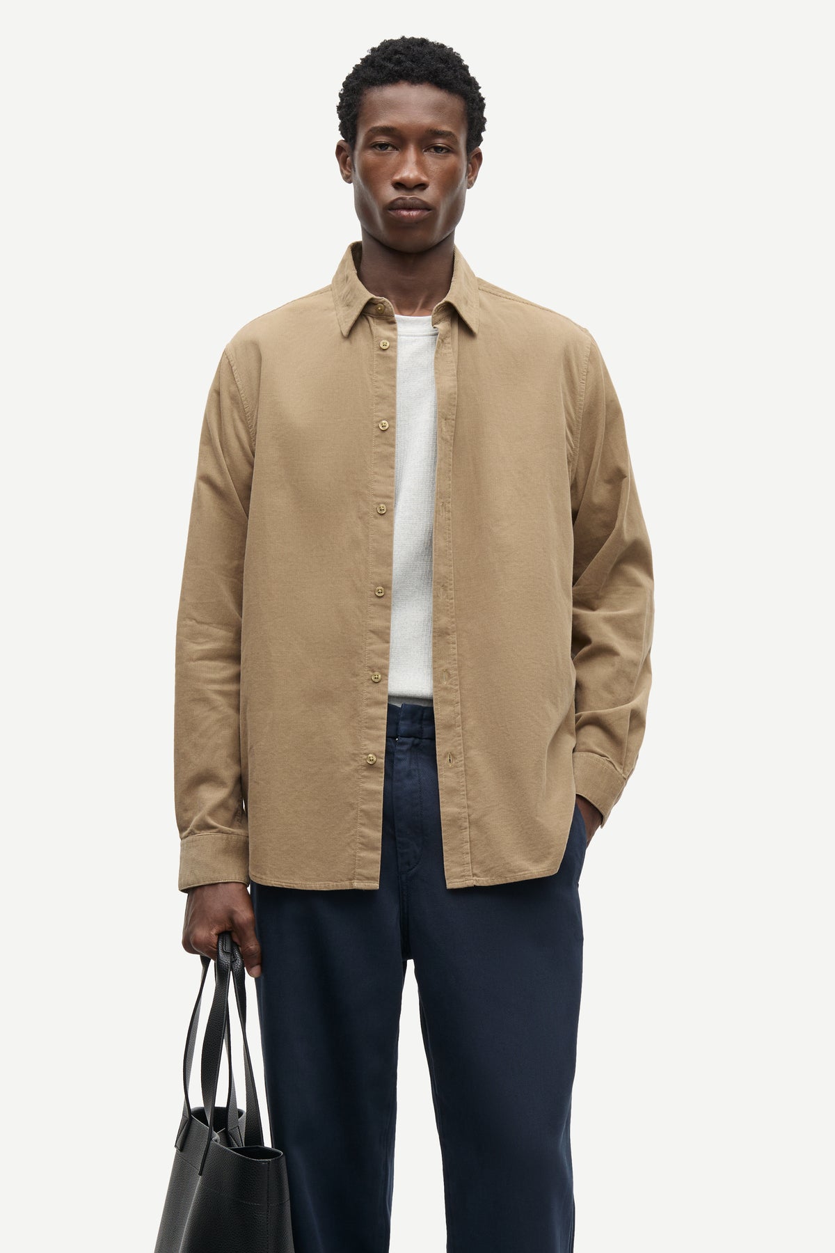 A man modeling a tan long-sleeve button-up shirt over a white t-shirt and navy blue trousers, holding a black tote bag against a light gray background.
