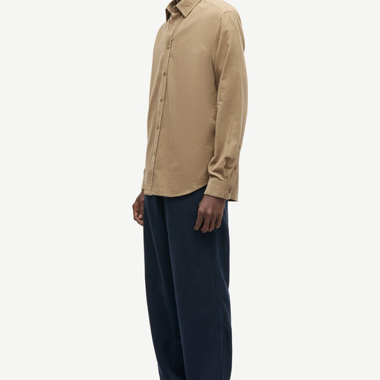 A man wearing a beige long-sleeve button-down shirt and dark navy trousers, standing in profile against a light gray studio background.