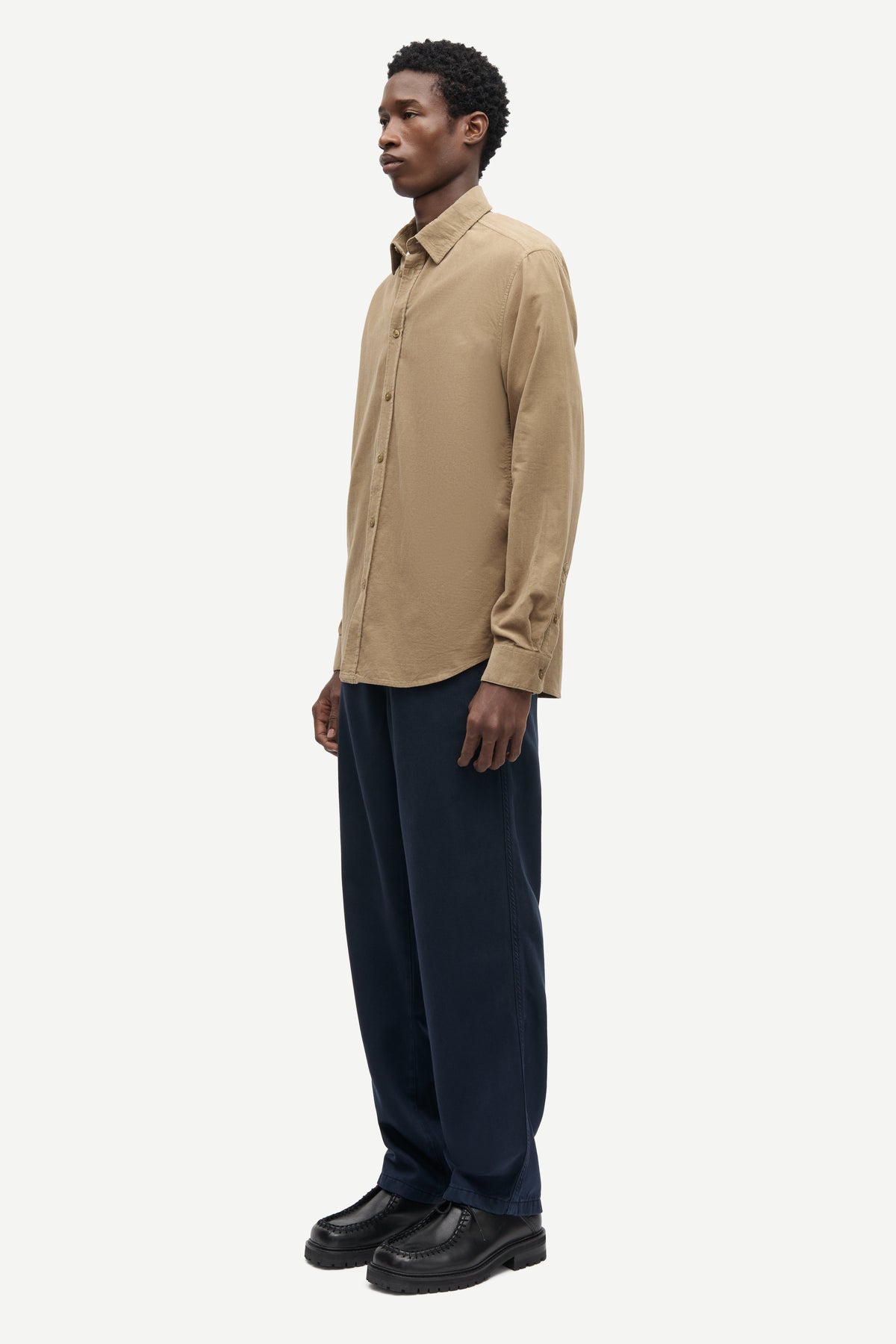 A man wearing a beige long-sleeve button-down shirt and dark navy trousers, standing in profile against a light gray studio background.