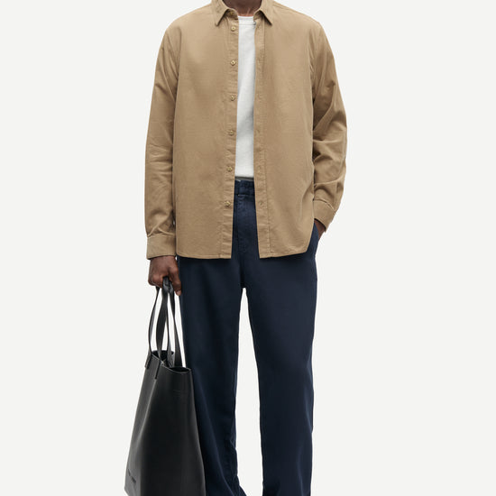 A man modeling a beige long-sleeve button-up shirt over a white t-shirt, paired with navy blue trousers and black shoes, holding a black tote bag against a light gray background.