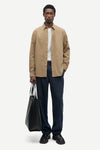 A man modeling a beige long-sleeve button-up shirt over a white t-shirt, paired with navy blue trousers and black shoes, holding a black tote bag against a light gray background.