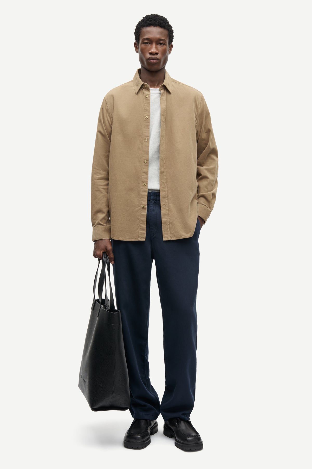 A man modeling a beige long-sleeve button-up shirt over a white t-shirt, paired with navy blue trousers and black shoes, holding a black tote bag against a light gray background.