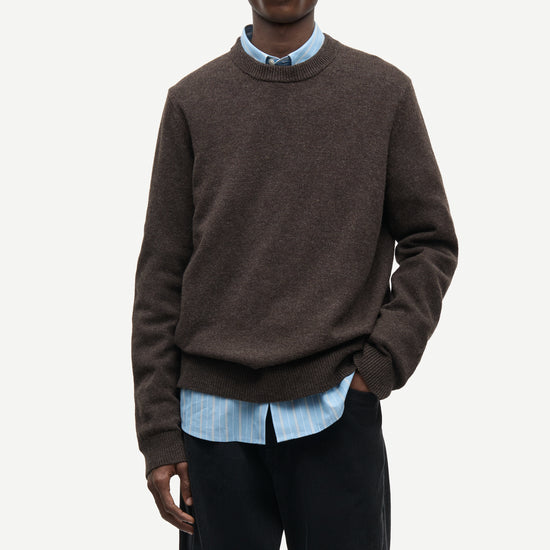A male model wearing a dark brown crewneck sweater over a light blue striped button-down shirt and black trousers, photographed against a clean off-white studio background.