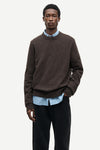 A male model wearing a dark brown crewneck sweater over a light blue striped button-down shirt and black trousers, photographed against a clean off-white studio background.