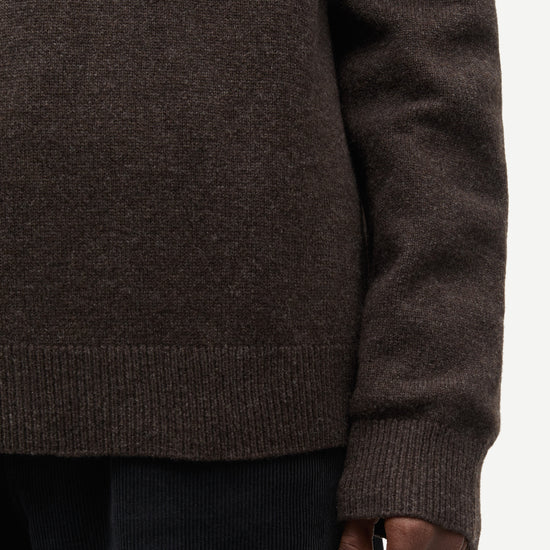 Close-up of a person wearing a dark brown knit sweater with ribbed cuffs and hem, paired with dark corduroy pants against a plain light background.