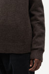Close-up of a person wearing a dark brown knit sweater with ribbed cuffs and hem, paired with dark corduroy pants against a plain light background.
