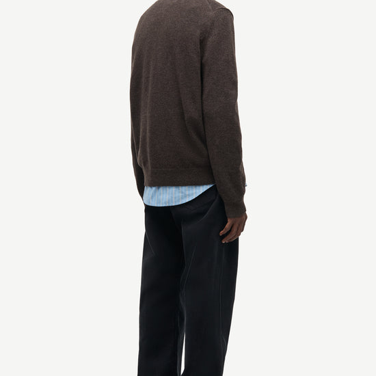 A man is shown from the back wearing a dark brown knit sweater, a light blue collared shirt, dark trousers, and dark loafers, against a plain light gray studio background.