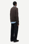 A man is shown from the back wearing a dark brown knit sweater, a light blue collared shirt, dark trousers, and dark loafers, against a plain light gray studio background.