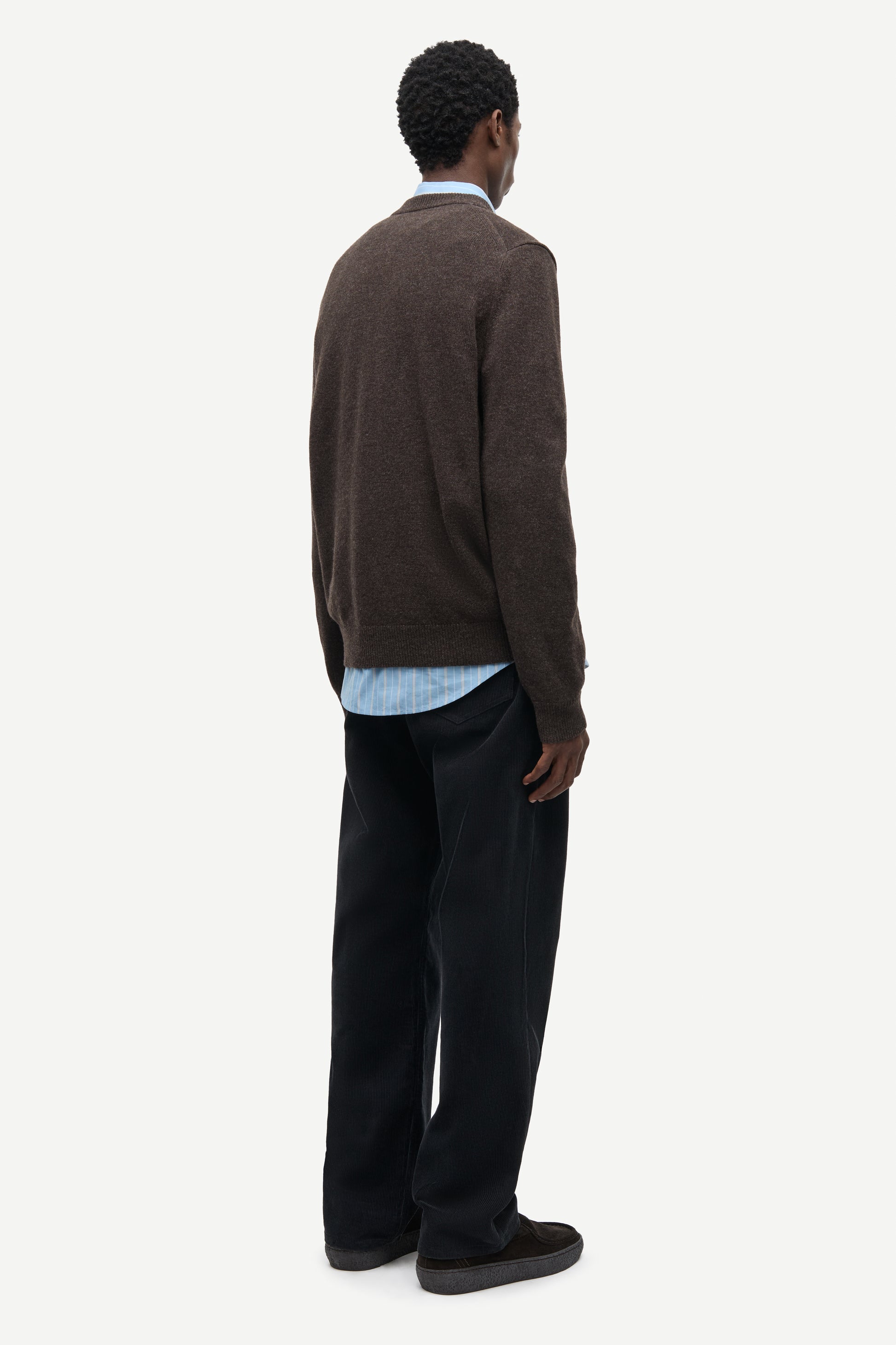 A man is shown from the back wearing a dark brown knit sweater, a light blue collared shirt, dark trousers, and dark loafers, against a plain light gray studio background.