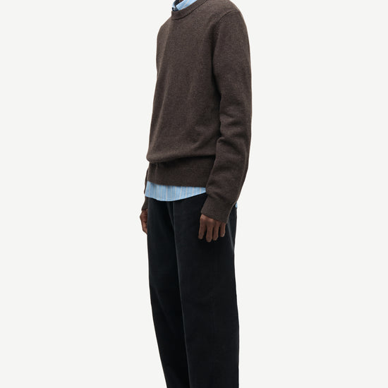A man wearing a brown knit sweater over a light blue collared shirt, paired with dark trousers and dark loafers, standing in profile against a plain light gray studio background.
