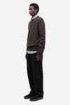 A man wearing a brown knit sweater over a light blue collared shirt, paired with dark trousers and dark loafers, standing in profile against a plain light gray studio background.