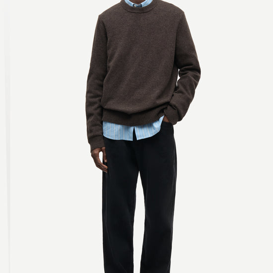 A man modeling a dark brown crewneck sweater layered over a light blue collared shirt, paired with dark trousers and dark brown suede shoes, against a plain light gray background.