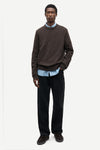 A man modeling a dark brown crewneck sweater layered over a light blue collared shirt, paired with dark trousers and dark brown suede shoes, against a plain light gray background.
