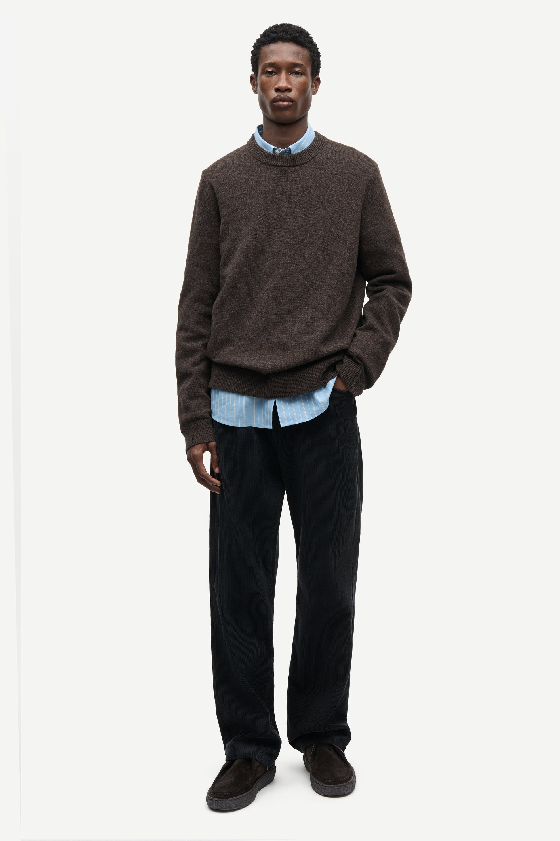 A man modeling a dark brown crewneck sweater layered over a light blue collared shirt, paired with dark trousers and dark brown suede shoes, against a plain light gray background.