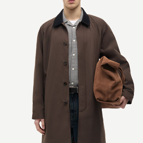 A male model wearing a dark brown mid-length coat over a checkered shirt and jeans, holding a matching brown suede-like pouch against a plain light gray studio background.