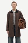 A male model wearing a dark brown mid-length coat over a checkered shirt and jeans, holding a matching brown suede-like pouch against a plain light gray studio background.