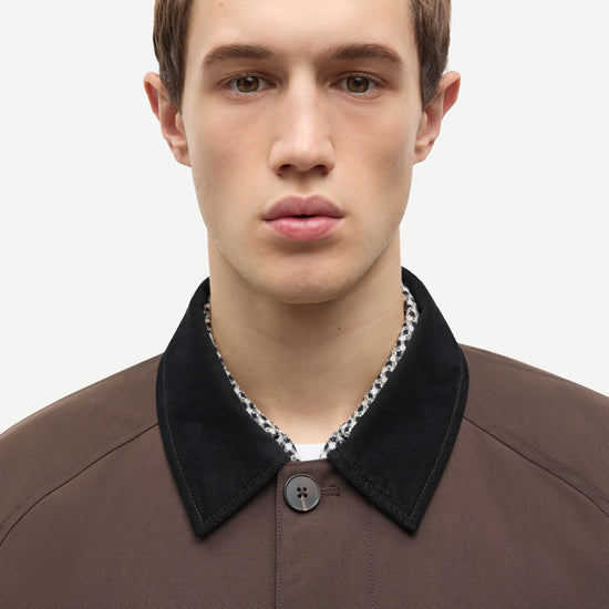 A close-up portrait of a male model wearing a brown textured jacket with a contrasting black velvet collar over a patterned shirt.