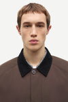 A close-up portrait of a male model wearing a brown textured jacket with a contrasting black velvet collar over a patterned shirt.