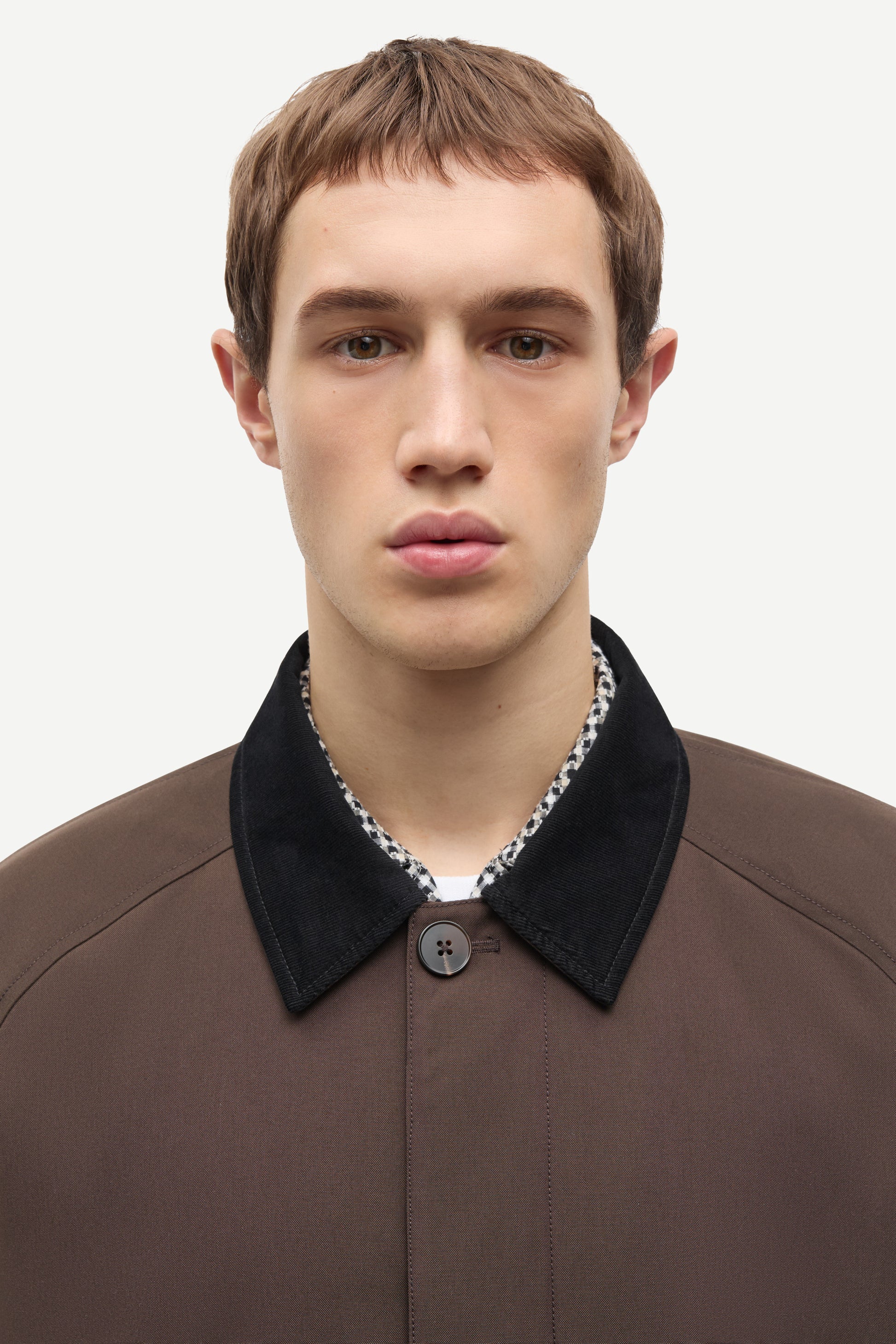 A close-up portrait of a male model wearing a brown textured jacket with a contrasting black velvet collar over a patterned shirt.