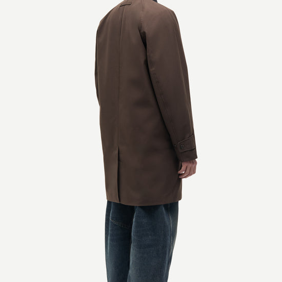 A man shown from the back wearing a dark brown mid-length coat over a black collared shirt, blue jeans, and black slip-on shoes, photographed against a plain light gray studio background.