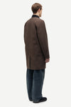 A man shown from the back wearing a dark brown mid-length coat over a black collared shirt, blue jeans, and black slip-on shoes, photographed against a plain light gray studio background.